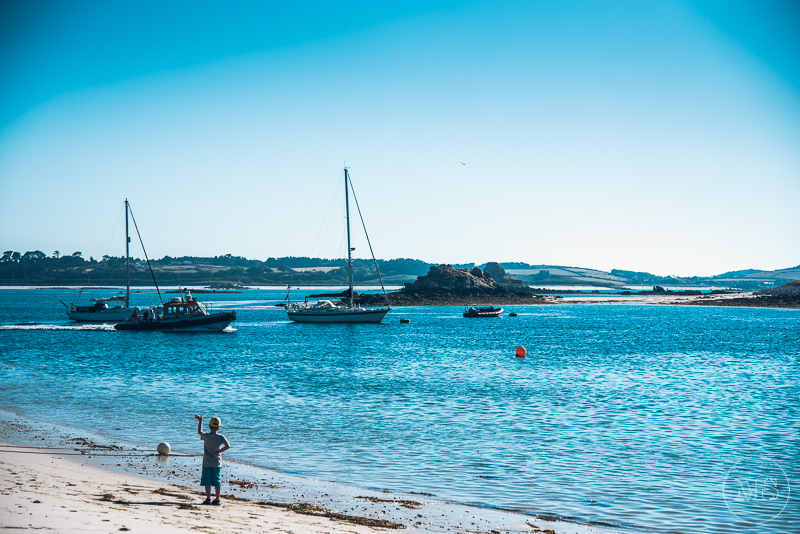 Child playing with a ball on a beach while sailboats float on calm water in the distance under a clear blue sky.