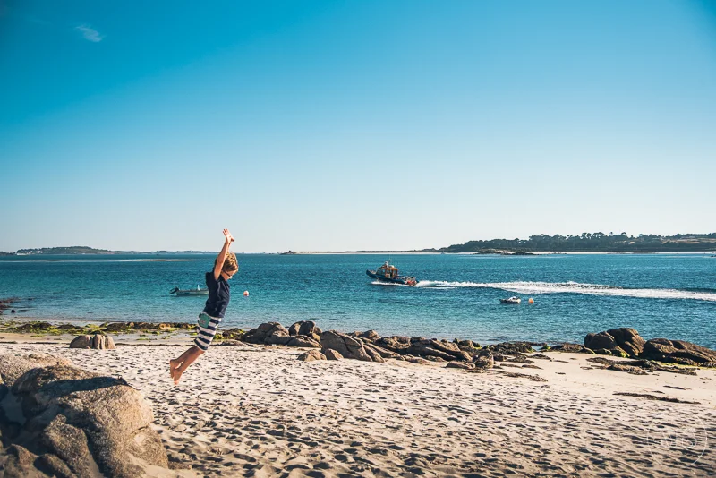 A child jumping on a sandy beach with rocks, a boat in the water, and a clear blue sky.