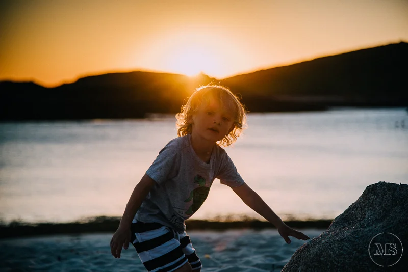 A young child with curly blonde hair standing near a body of water at sunset, with hills or mountains in the background.