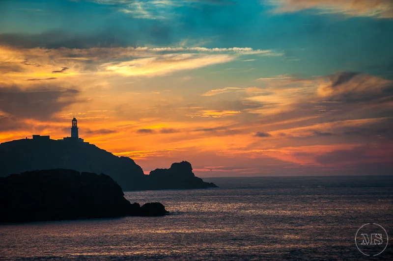 Sunset over the ocean with a lighthouse on a hill in the distance.