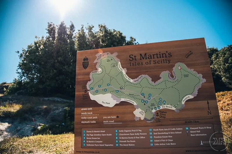 A wooden map sign of St. Martin's Isles of Scilly showing walking trails and points of interest, with logos and a compass in the corners, set outdoors with trees and a blue sky.