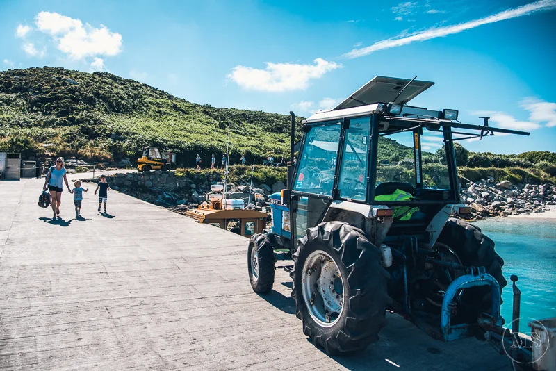 A tractor parked on a concrete dock next to the water, with a woman and three children walking hand-in-hand on the dock. In the background, there are hills, a few other vehicles, and people near the shoreline.