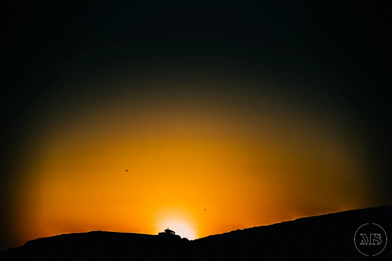 Isles of scilly photography. Sunset over a mountain ridge with a silhouette of a small structure or house on the peak, early evening sky with orange and black colors.