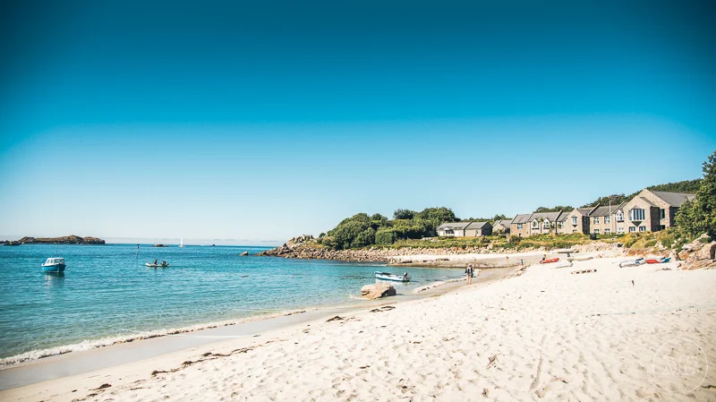 Isles of scilly photography. Beach scene with white sand, boats, and houses in the background under a clear blue sky.