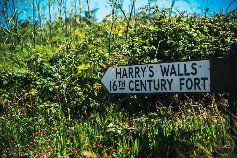 Isles of scilly photography. A black and white directional sign reading "Harry's Walls 16th Century Fort" pointing left, situated among green foliage with a blue sky background.