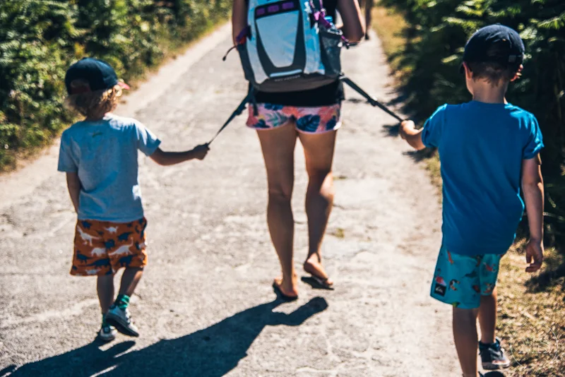 A woman walking with two young boys on a dirt path in a sunny outdoor setting.