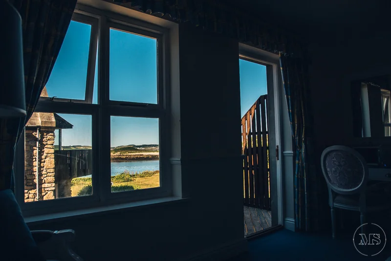 View through large window of a seaside landscape with water, land, and sky; partial door open to balcony or patio with wooden railing; interior includes curtains and a white chair.