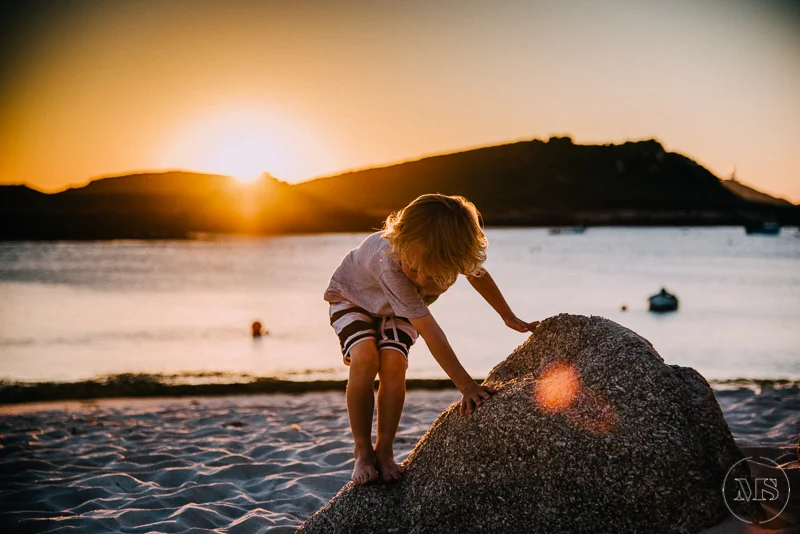 Isles of scilly photography. A young boy climbing on a large rock on a beach during sunset, with a body of water and hills in the background.
