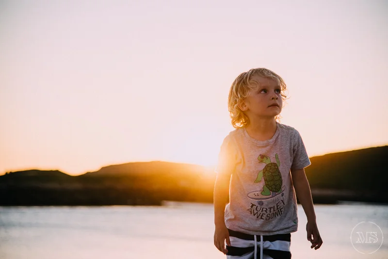 A young boy with curly blond hair standing outdoors during sunset, with a river and rolling hills in the background, wearing a gray t-shirt with a turtle graphic.