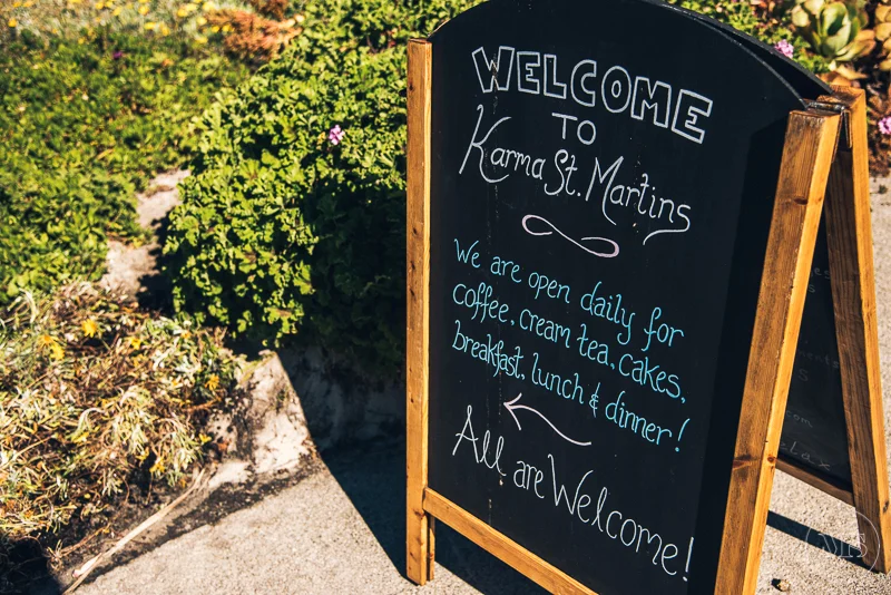 A black chalkboard sign on a wooden stand with white and blue handwritten text welcoming visitors to Karma St. Martins, noting the open daily hours and offerings like coffee, cream tea, cakes, breakfast, lunch, and dinner, placed outdoors among plant