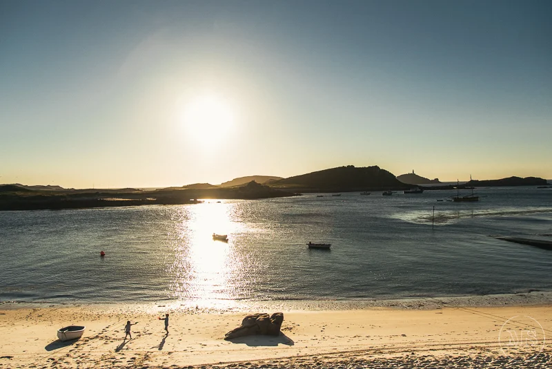 Isles of scilly photography. Sunset over a calm body of water with boats floating near the shore, sandy beach in the foreground, and a hilly landscape in the distance.