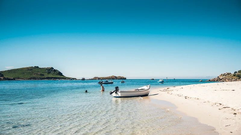 A peaceful beach with white sand, a small boat in shallow water, two people swimming, and several boats anchored farther out. There are green hills in the background under a clear blue sky - Isles of Scilly Professional Photographer