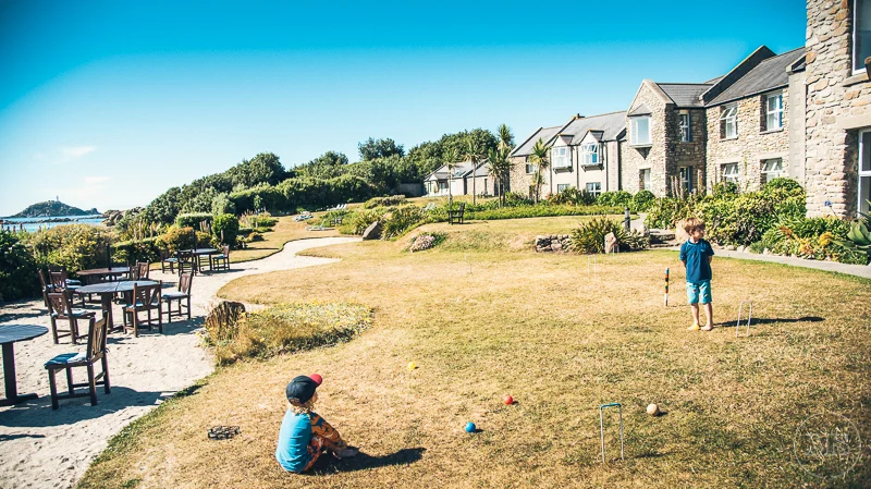 Isles of scilly photography. Two children playing lawn games on a grassy yard near stone houses on a sunny day.