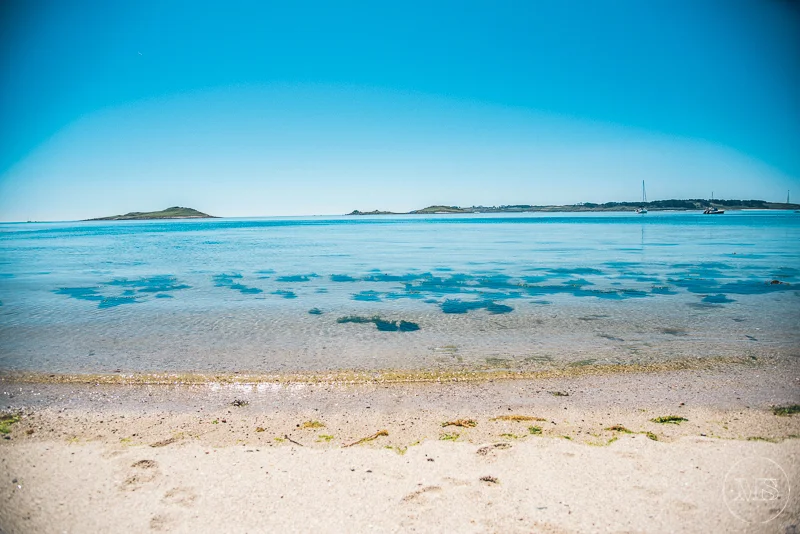 Isles of scilly photography. Calm beach with clear shallow water, sandy shore with seaweed, distant islands, and sailboats on the horizon under a blue sky.