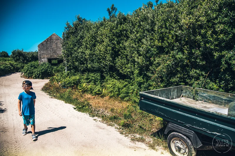 A young boy wearing a blue shirt, blue shorts, and sunglasses walking along a dirt path near green bushes and trees, with a trailer attached to a vehicle on the right side and a rustic stone building in the background on a sunny day.