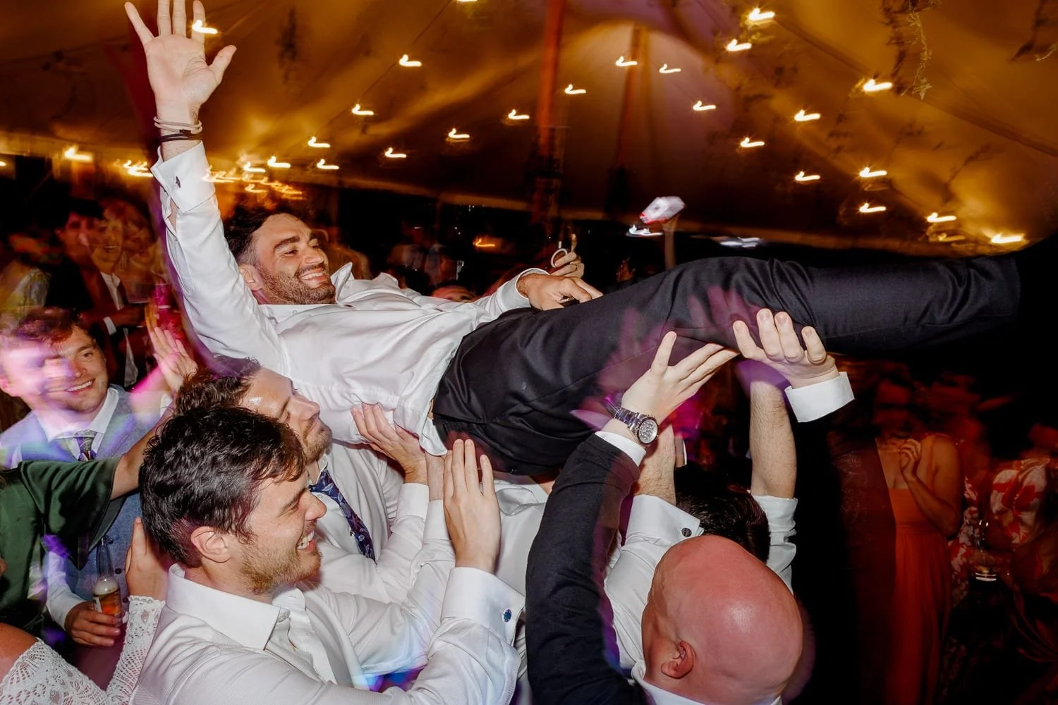 A man being lifted horizontally by a group of people at a celebration or party, smiling and waving with his right hand, with a festive atmosphere and colorful lights in the background.