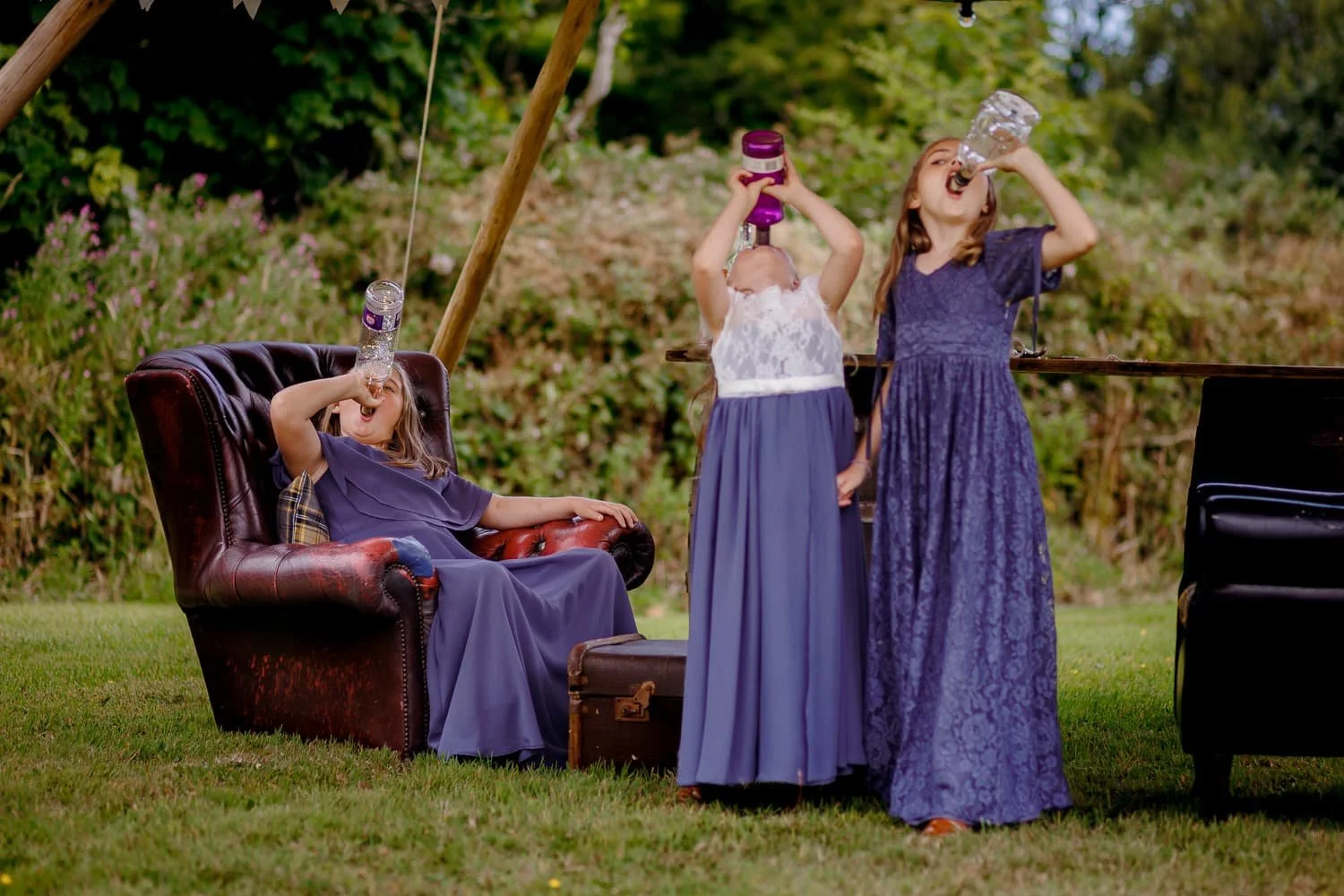 Three children in blue dresses drinking water outdoors, one sitting on a vintage armchair and two standing, with greenery in the background.