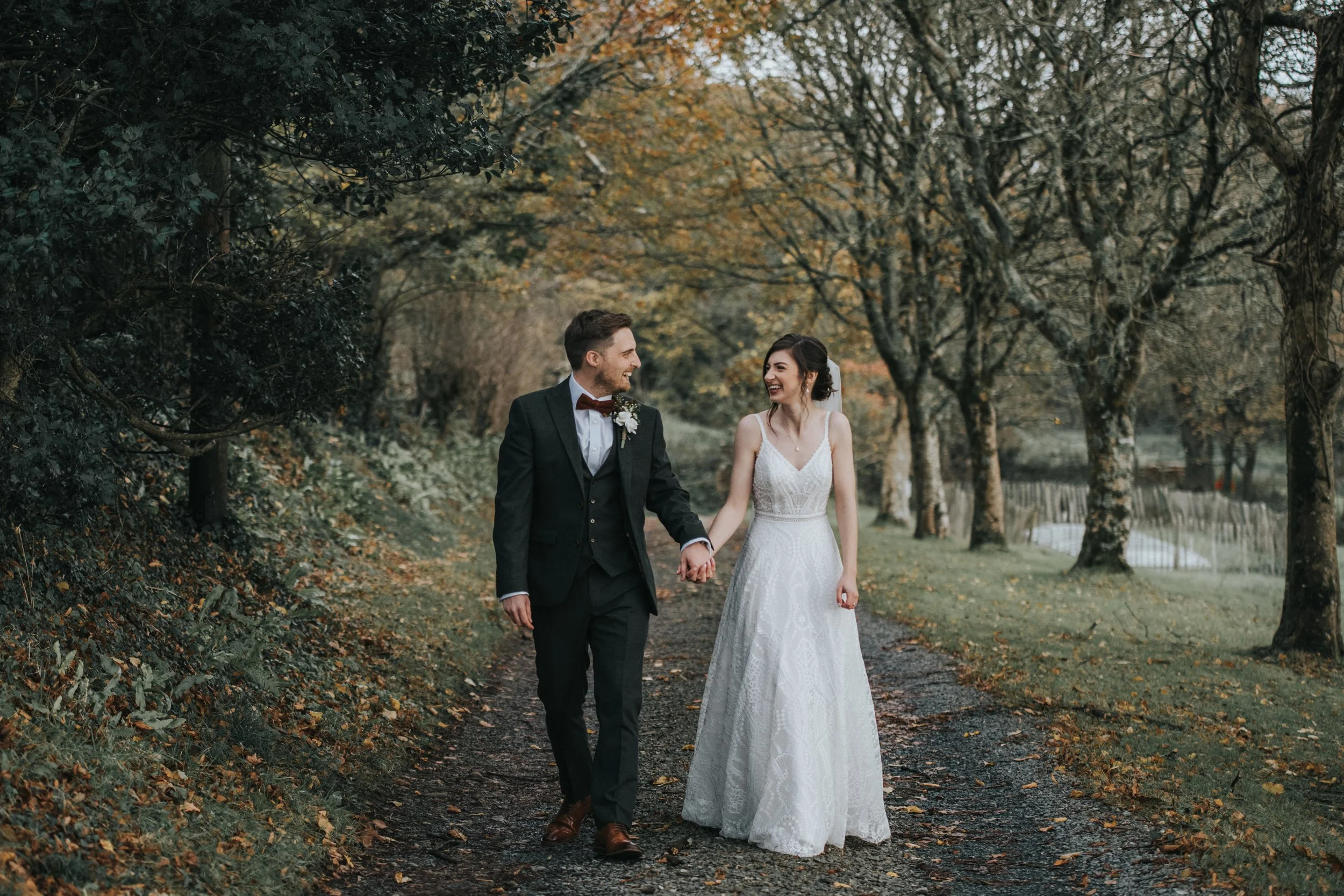 A newlywed couple walking hand in hand on a dirt path in a park during autumn, surrounded by trees with fall foliage.