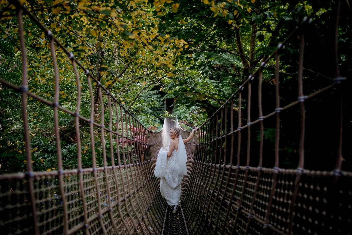 A woman in a white wedding dress and veil standing on a hanging bridge in a green, wooded area, holding onto the railings and looking back.