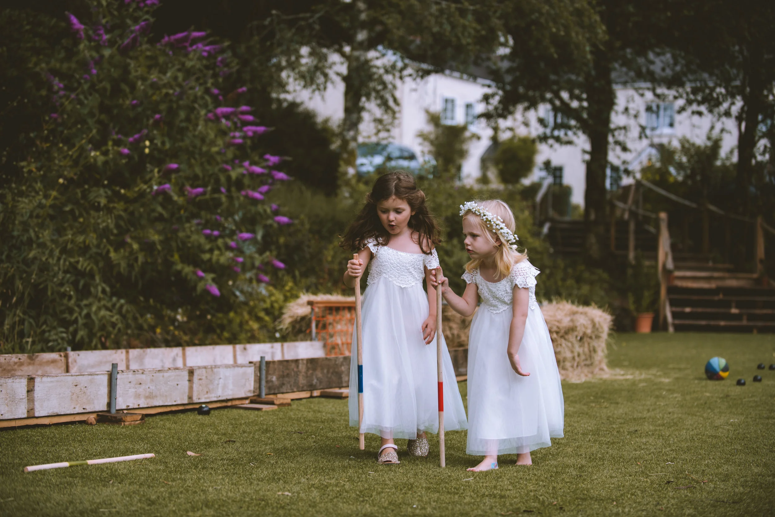 Two young girls dressed in white dresses are playing mini golf outdoors on a grassy area, one girl supporting the other as they hold golf clubs.