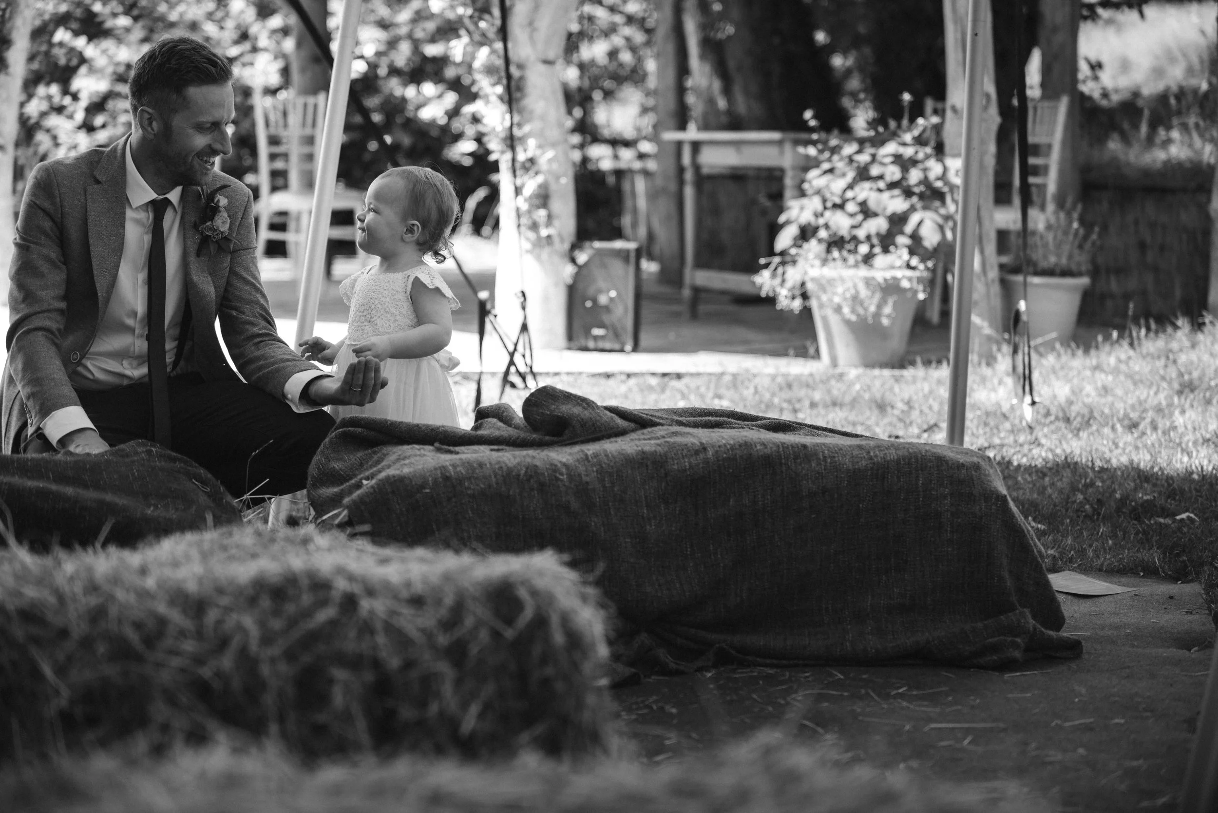 A man in a suit is sitting outside on a blanket with a young girl in a dress, both smiling and looking at each other. It appears to be at an outdoor event or gathering.