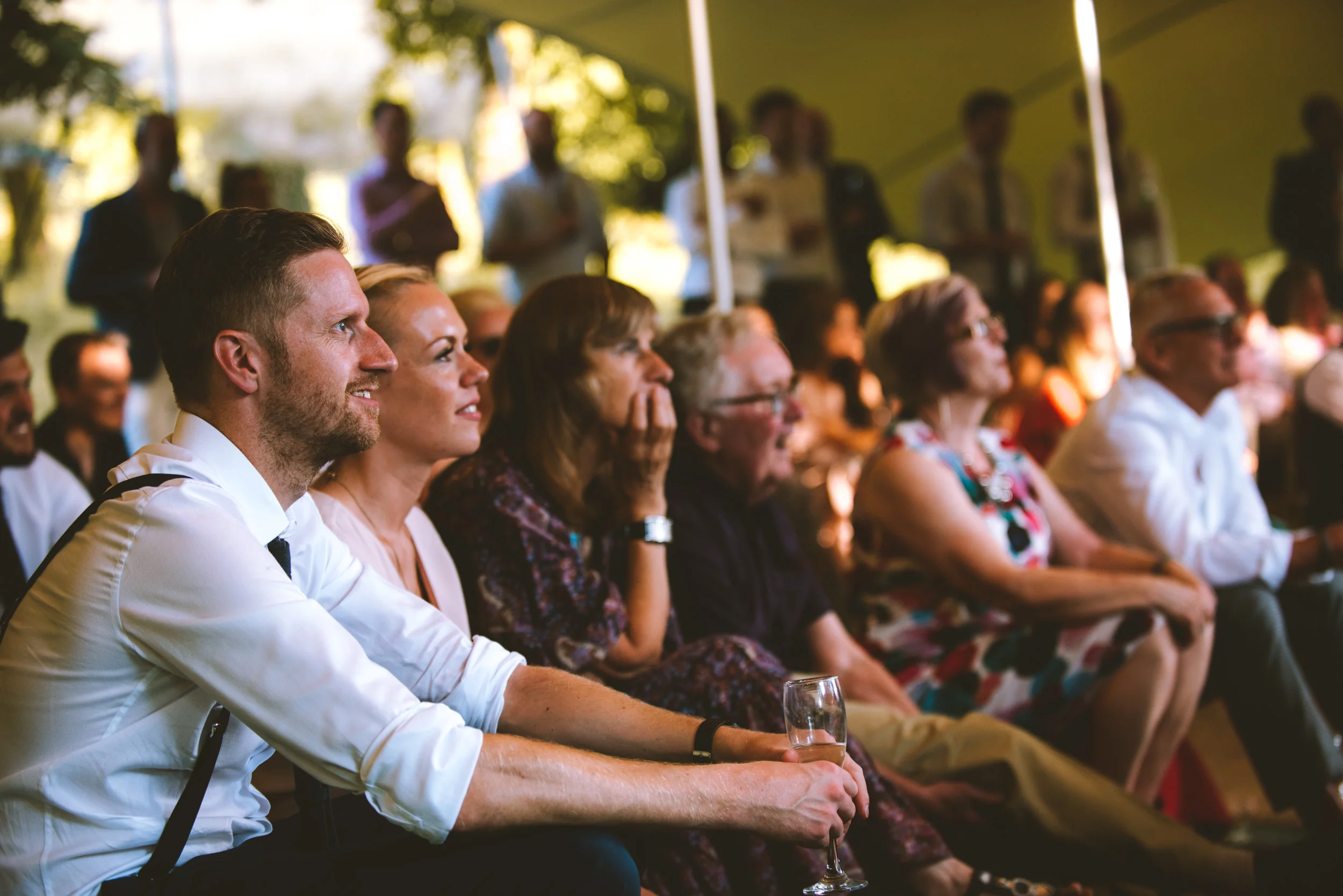 Audience seated and watching at an outdoor event under a tent, with some individuals smiling and looking attentively.