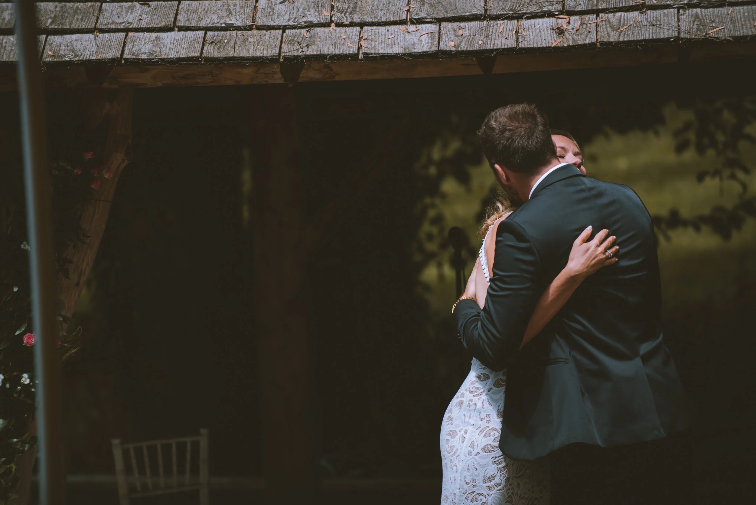 A couple in formal attire dancing closely with their arms wrapped around each other, in an outdoor setting under a wooden roof structure, during the evening or night with soft lighting.