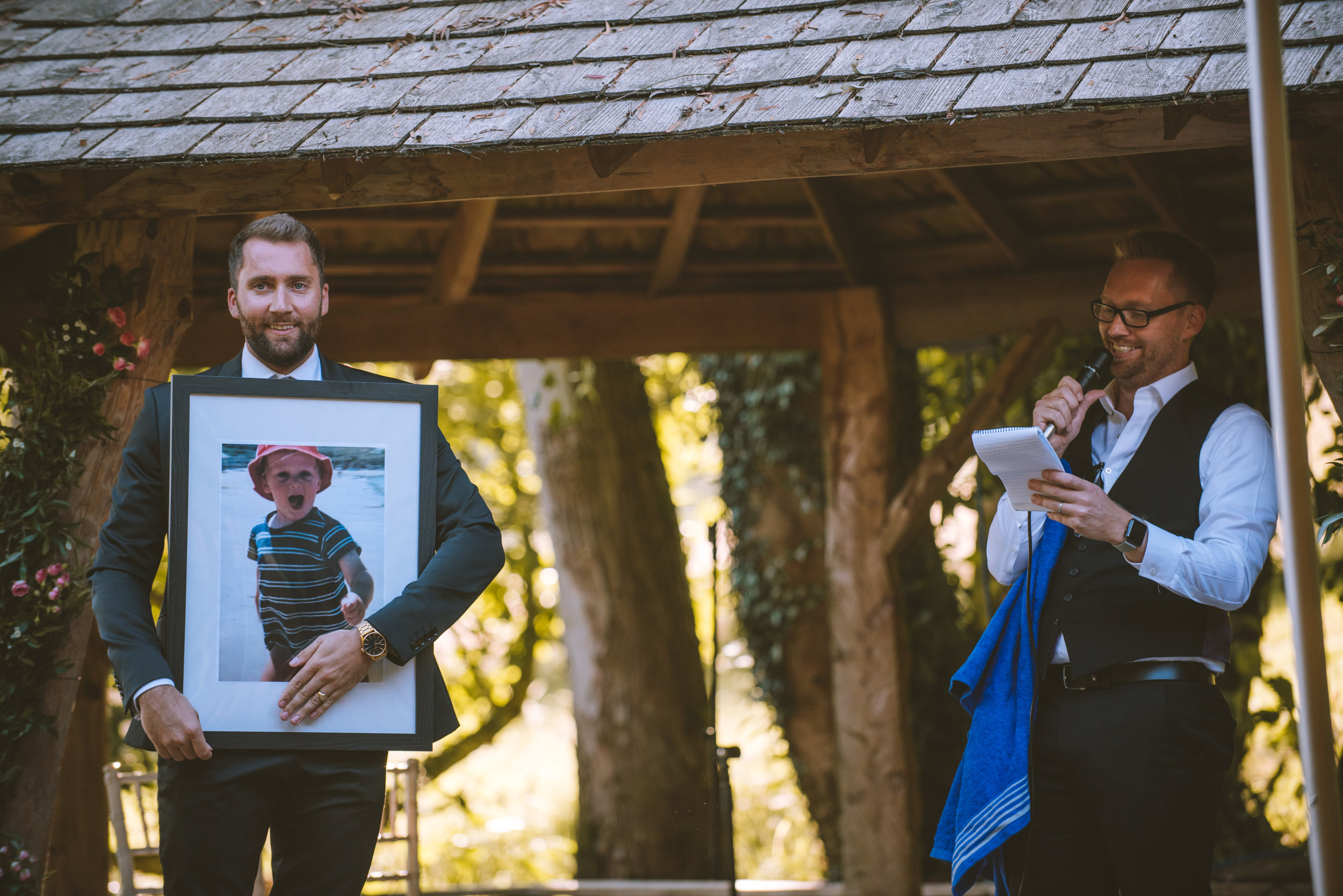 A man in a suit holds a large framed photo of a young boy wearing a striped shirt and red hat, standing outdoors. Another man, dressed in a vest and white shirt, is speaking into a microphone and holding a notepad, standing under a wooden gazebo with