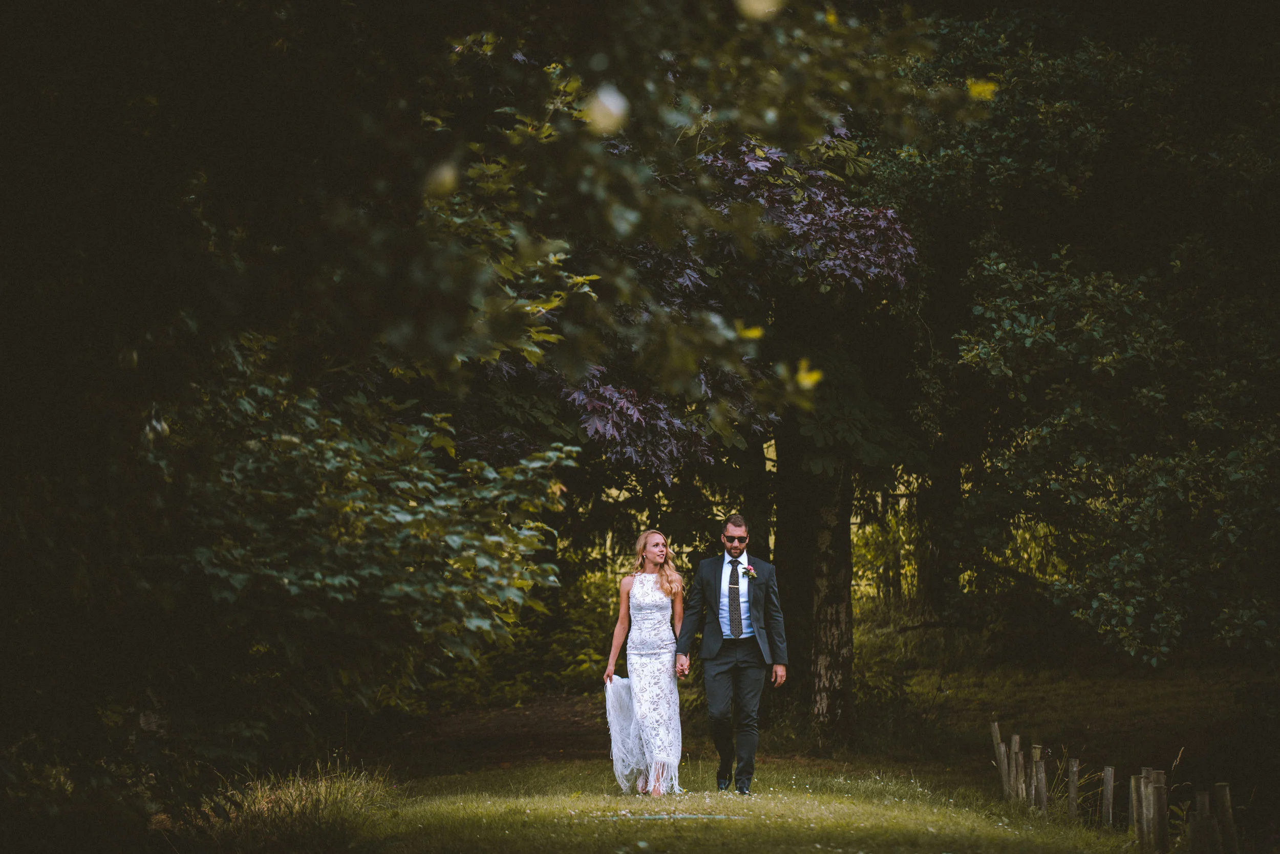 A bride and groom walking hand in hand through a lush green outdoor area during sunset or late afternoon, surrounded by trees and foliage.