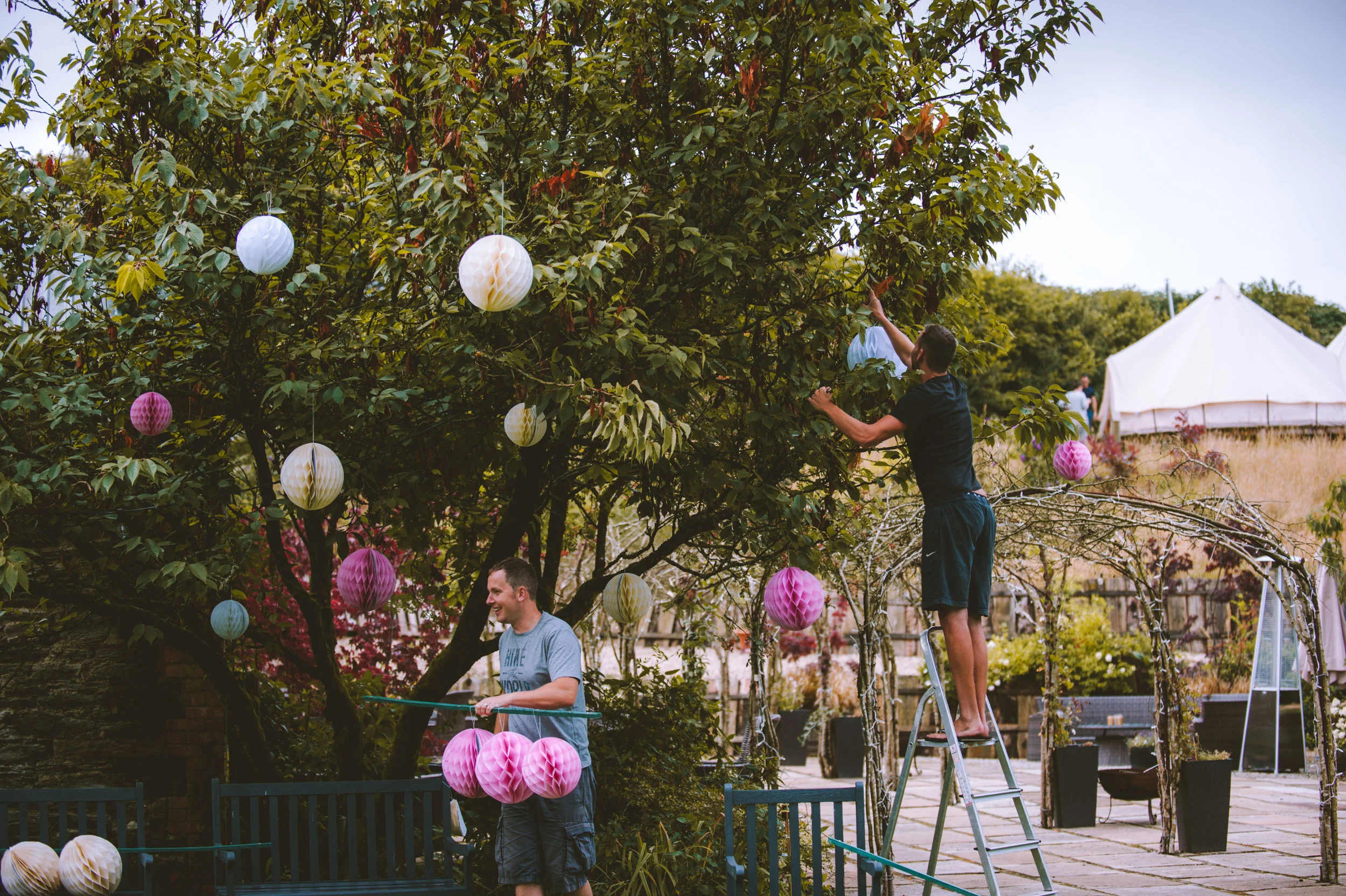 Two men decorating a garden with colorful paper lanterns and hanging lights, one on a ladder hanging lanterns from a tree, the other standing by the bench holding lanterns.