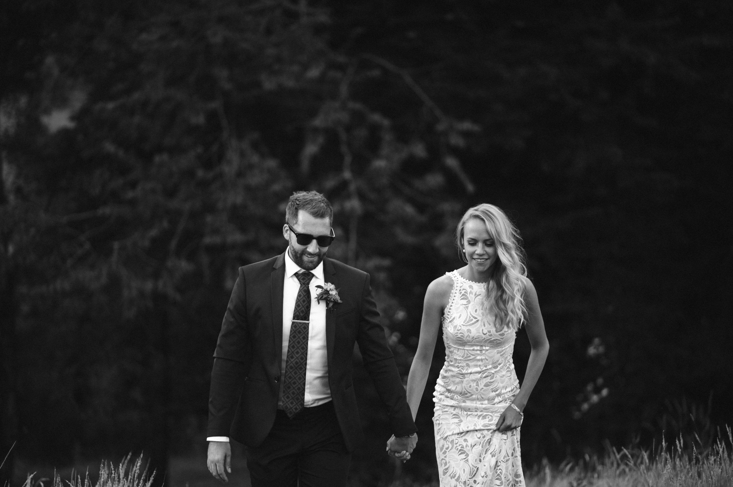 Black and white photo of a couple holding hands, walking outdoors in a natural setting, dressed in wedding attire. The man wears sunglasses, a suit, and a tie, while the woman is in a lace wedding dress with long wavy hair.
