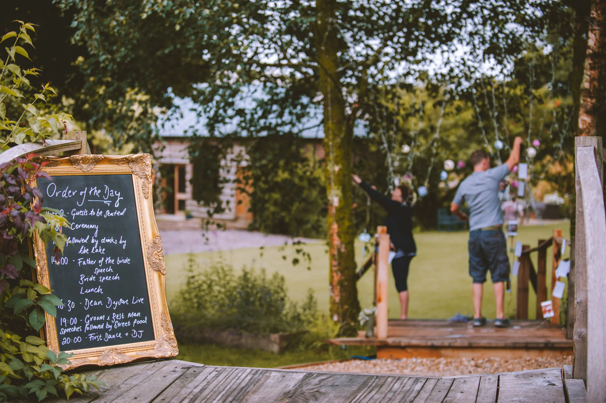 A decorated outdoor wedding venue with a path leading to a small wooden bridge, a blackboard with event schedule in an ornate gold frame, and a man and woman in the background near hanging paper or fabric decorations under trees.