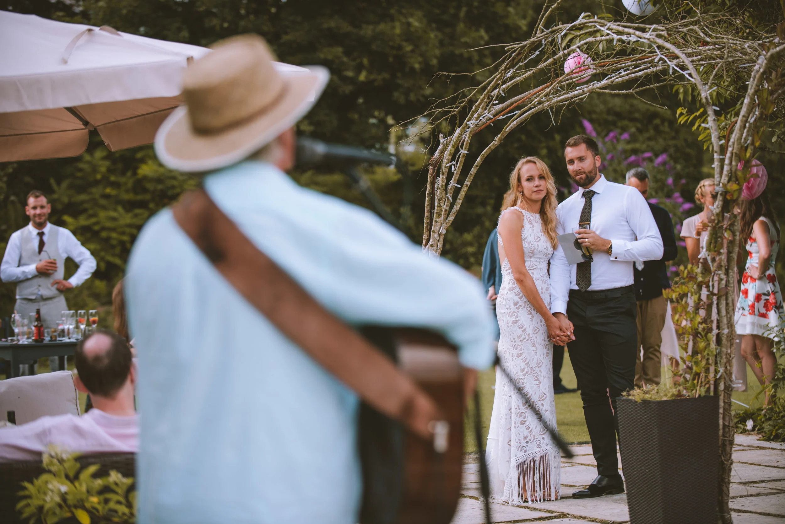 A couple holding hands at a wedding ceremony outdoors, surrounded by guests and decorative arch, while a musician plays guitar in the foreground.