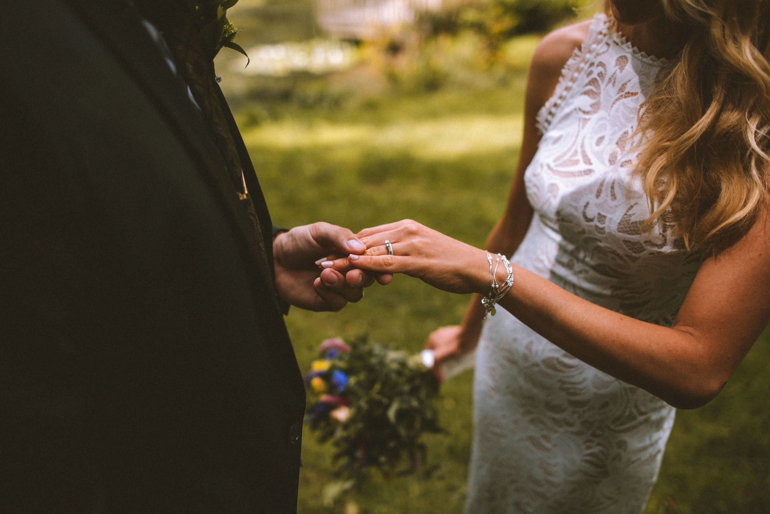 A couple at a wedding holding hands. The bride is wearing jewelry and a lace dress, and the groom is dressed in a dark suit. The bride is playing with the groom's wedding ring with her left hand, which has a ring on her finger. The bride's bouquet is