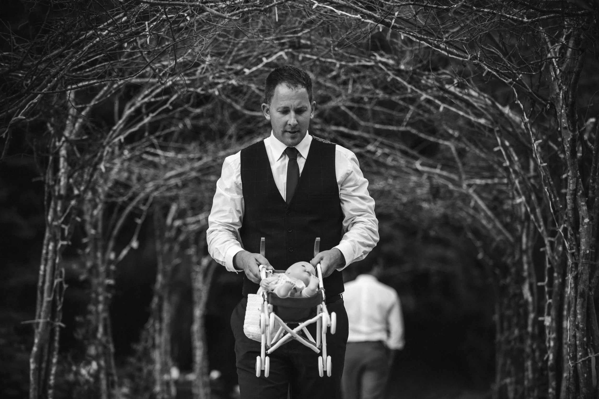 A man in a vest and tie pushing a doll stroller with a doll inside, walking through a tree-lined tunnel, in black and white.