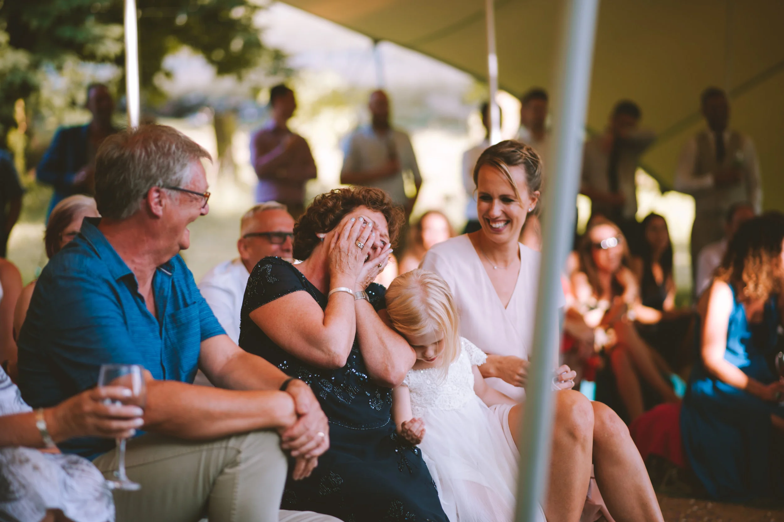 Group of people laughing and smiling at an outdoor event under a tent, with some standing and some seated.