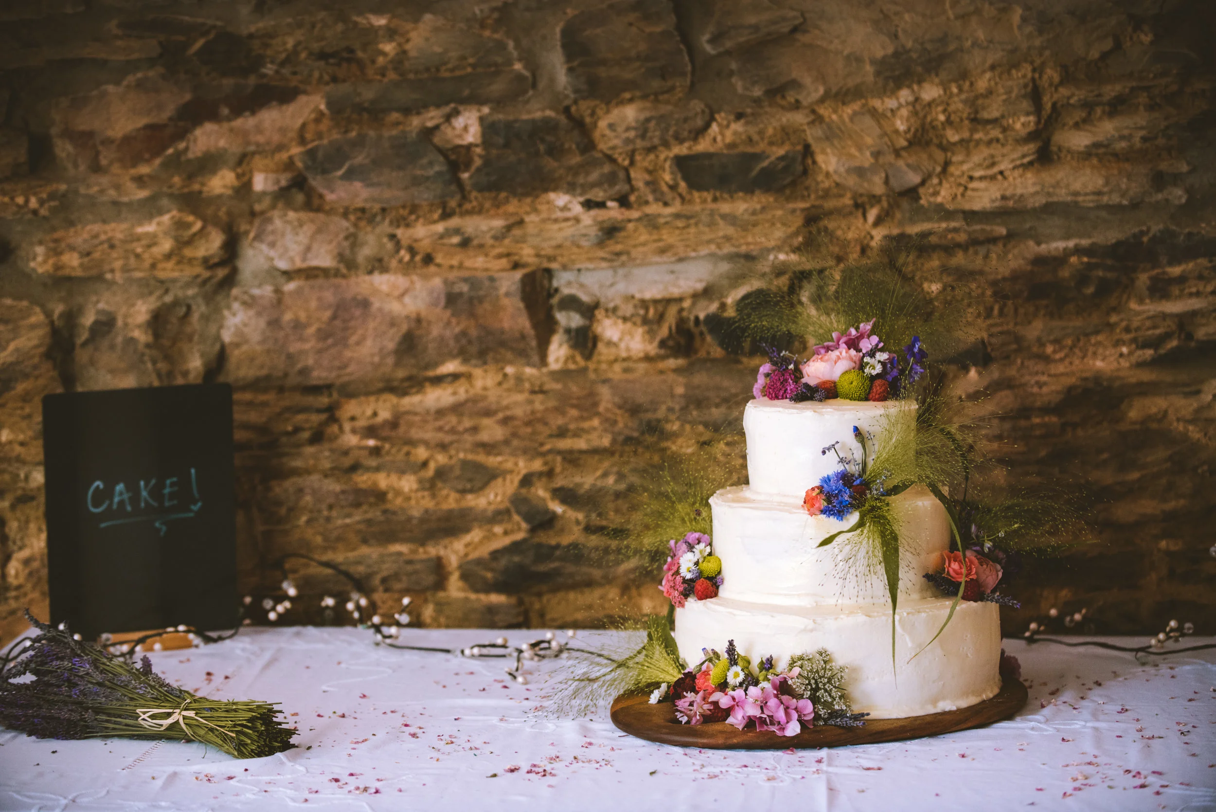 Three-tiered white wedding cake decorated with colorful flowers and greenery, placed on a wooden base on a white tablecloth with scattered confetti; a bouquet of purple flowers and a black chalkboard sign with the word 'CAKE!' written in blue, agains