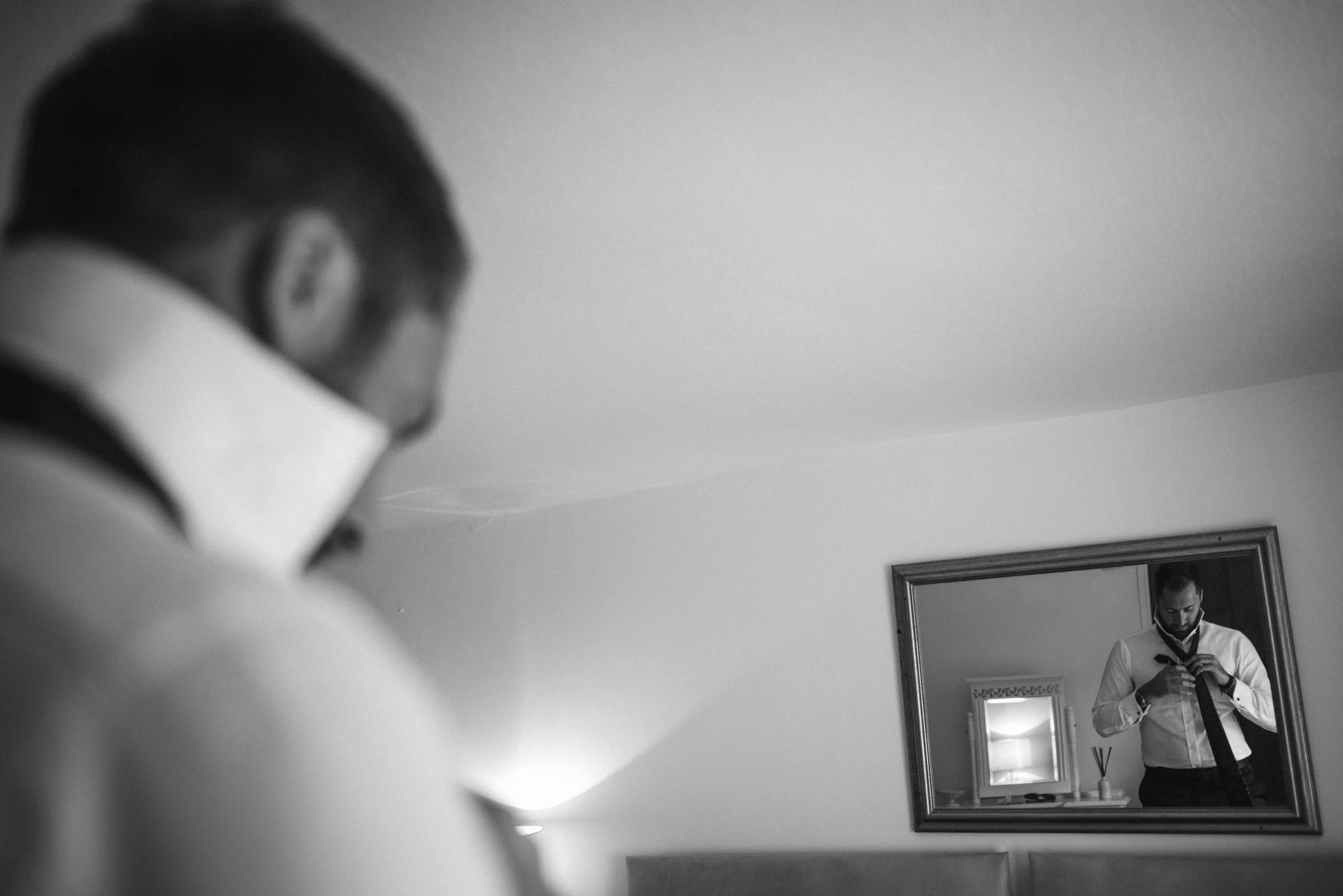 A man adjusting his tie in front of a mirror, with a second, blurred image of himself visible in the mirror and a third, larger mirror on the wall reflecting him.