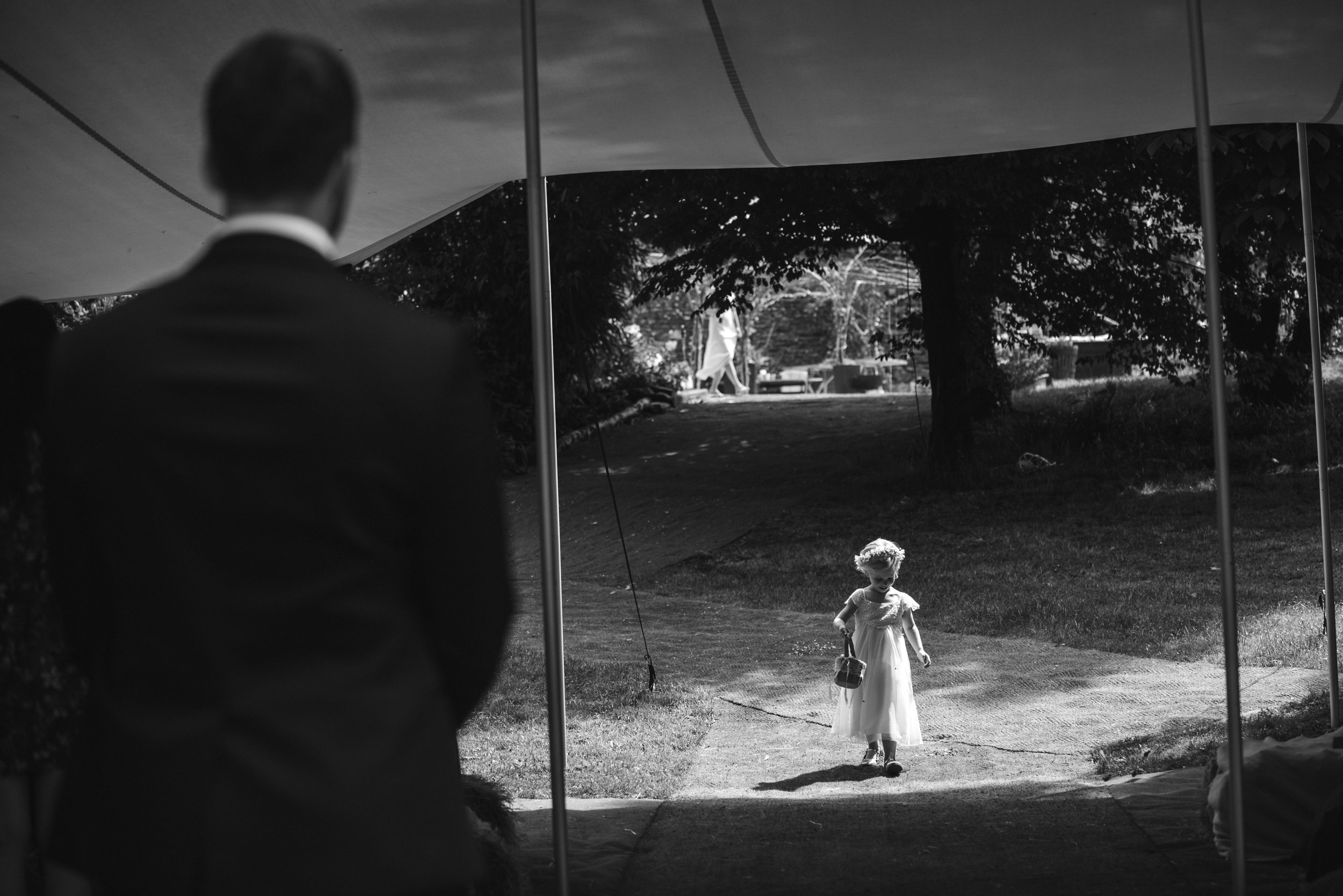 A young girl in a dress and crown carrying a basket walking on a path outside, seen from behind a tent or canopy. A man in a suit stands nearby, partially visible from behind, with trees and other people in the background.