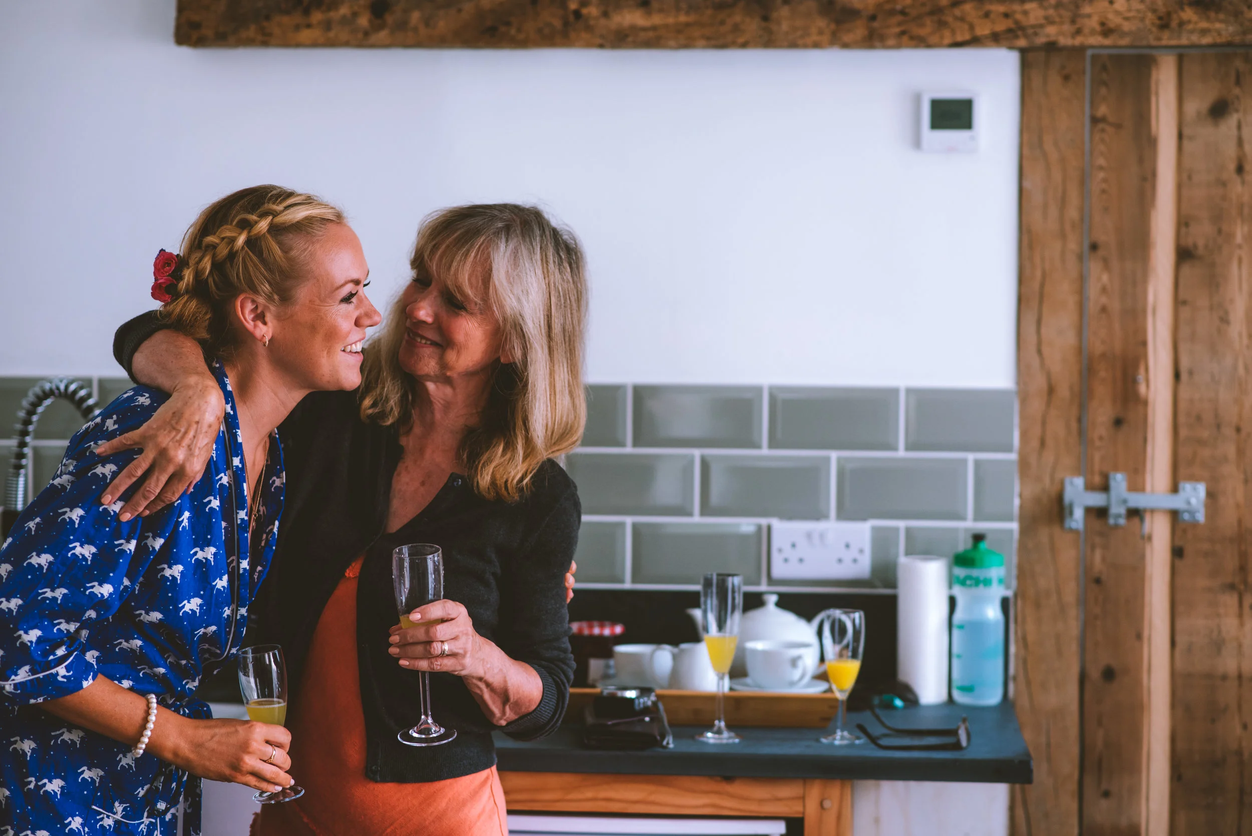 Two women are embracing and smiling at each other in a kitchen, holding champagne flutes with orange juice. The kitchen has a black countertop, gray tiles, and wooden accents.