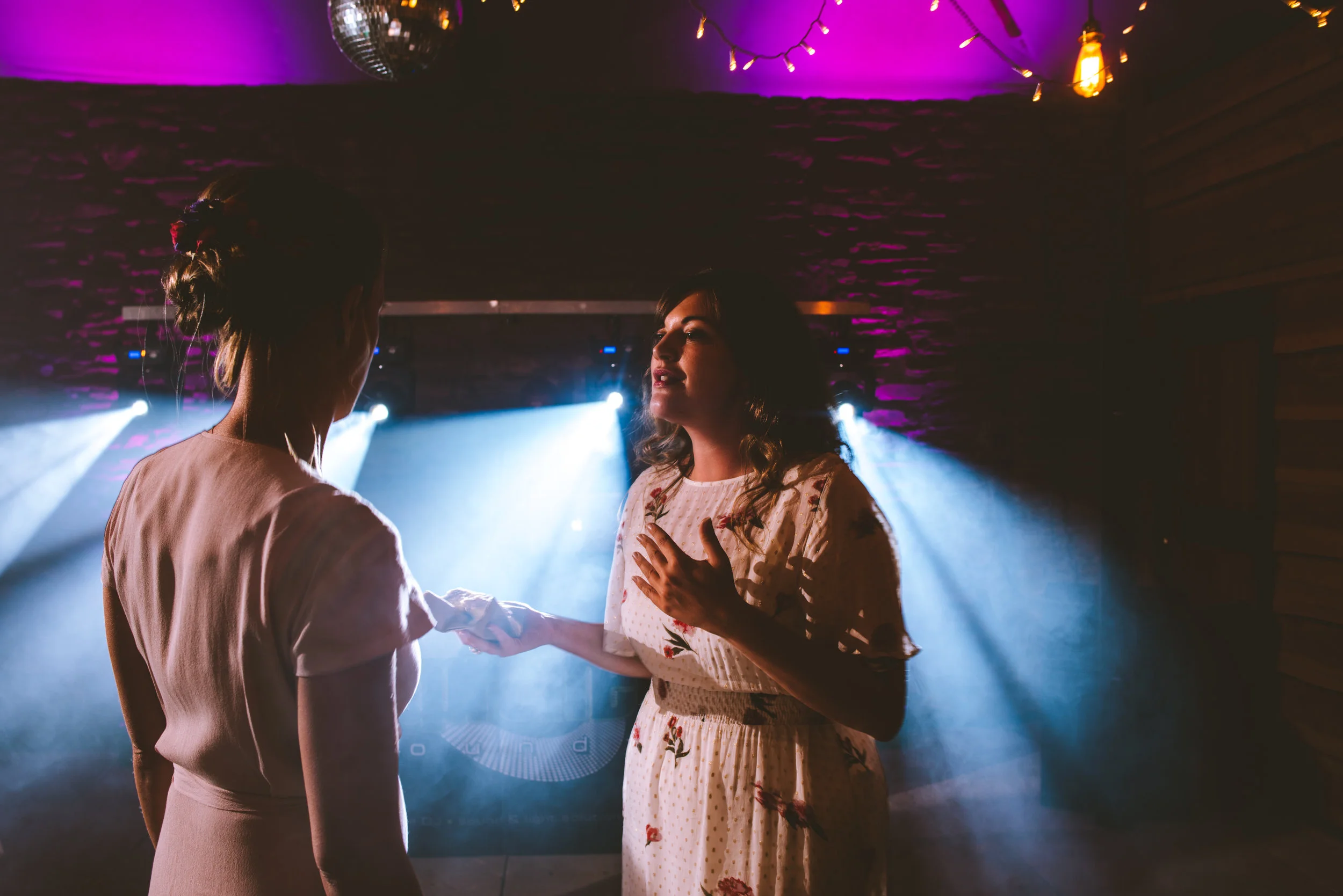 Two women are dancing at a party or dance club. The woman in the foreground has her back to the camera and is wearing a pale pink dress. The woman facing the camera is wearing a light-colored, floral dress with her hand on her chest, and they are hol