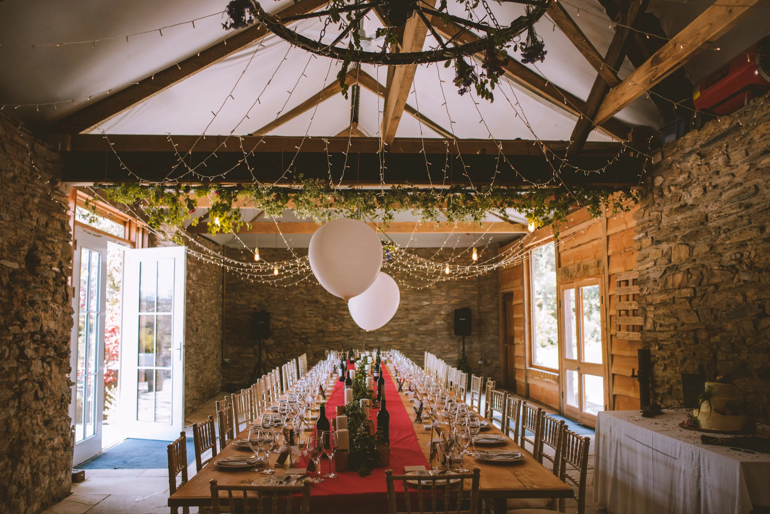 Long dining table set for a celebration in a rustic barn, decorated with string lights, large hanging balloons, and greenery, with stone and wood walls and windows letting in natural light.