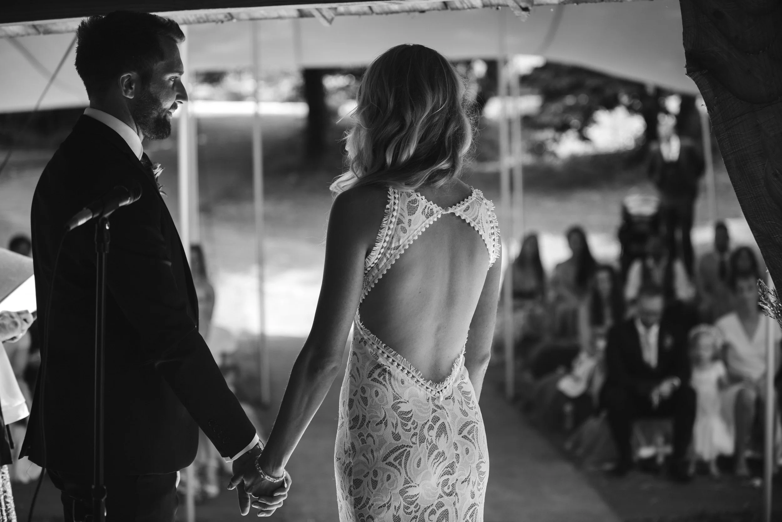 A bride and groom holding hands during their outdoor wedding ceremony, with guests seated in the background.