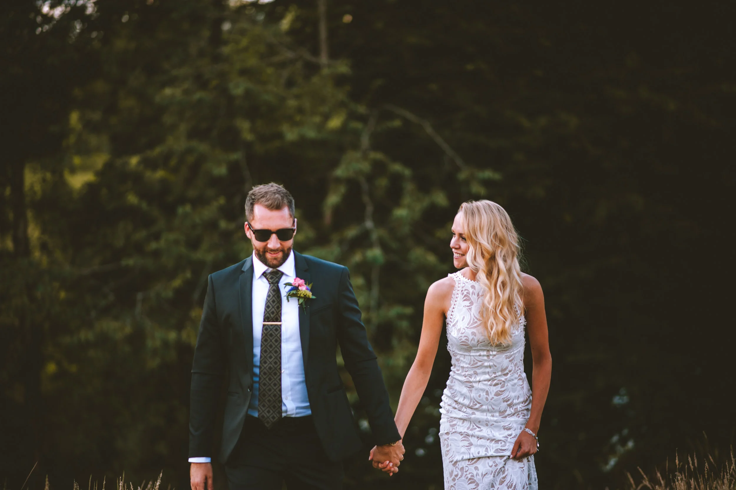 A couple dressed in wedding attire holding hands outdoors, with the groom in a dark suit and the bride in a white lace dress, smiling at each other against a backdrop of trees.