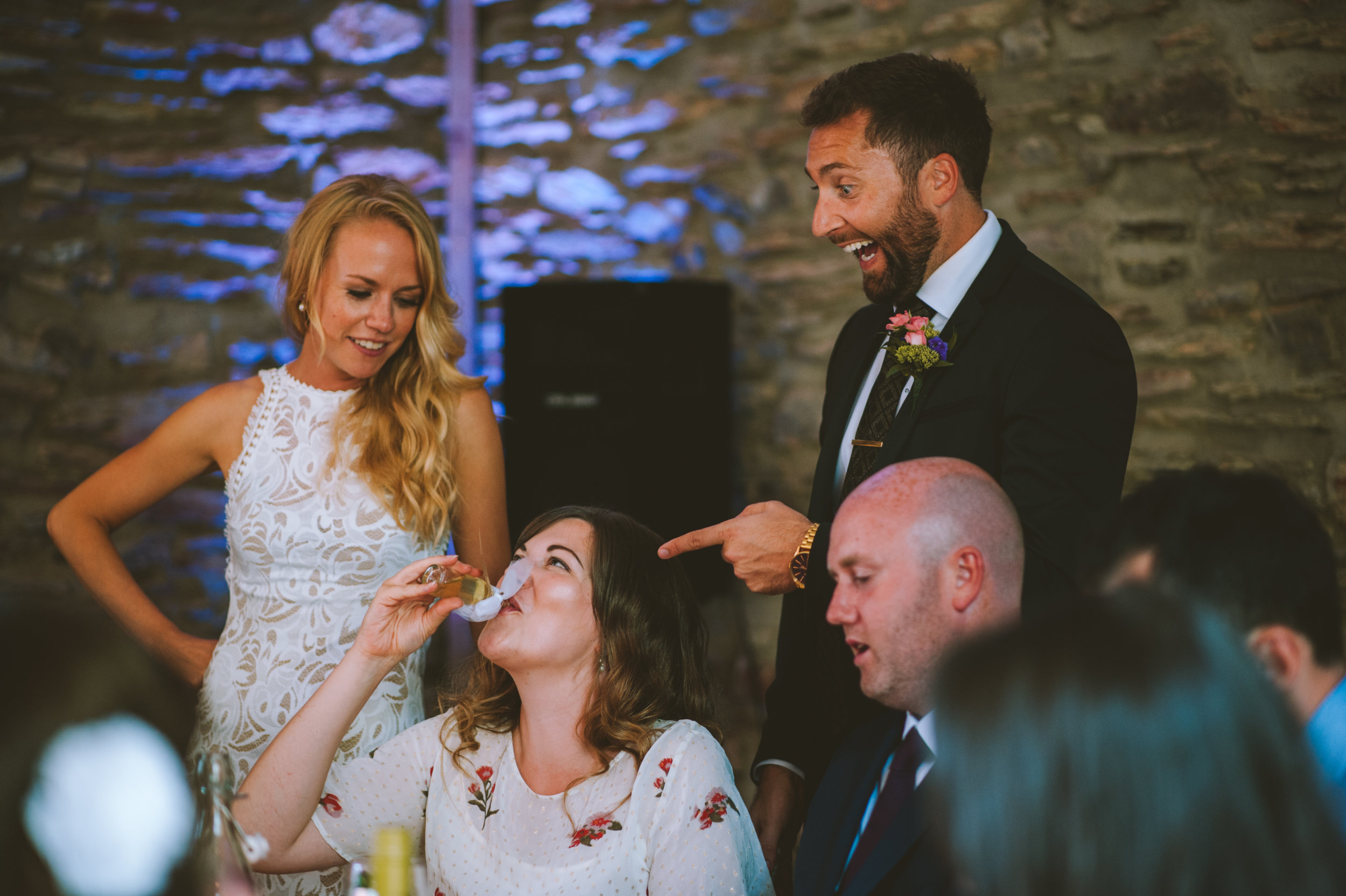 People celebrating at a wedding reception, one woman is drinking from a glass and smiling, a man in a suit with a boutonnière pointing at her, a woman with blonde hair looking on, and others in the background.