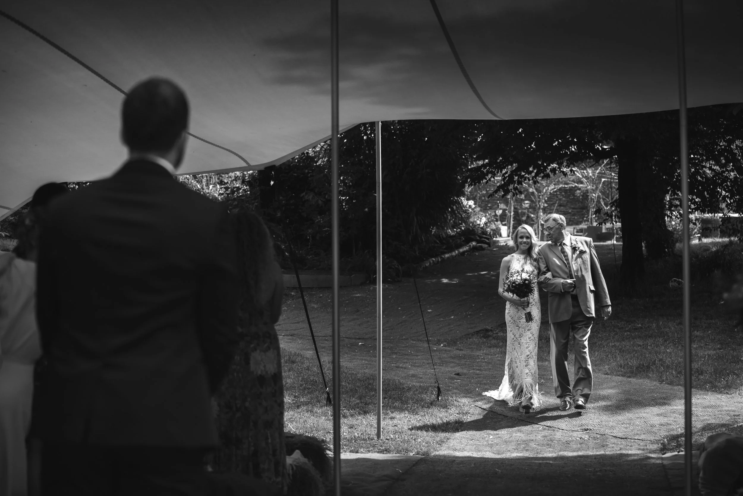 A bride walking with an older man, possibly her father, at her wedding, outdoors under a large canopy with guests watching in the background.