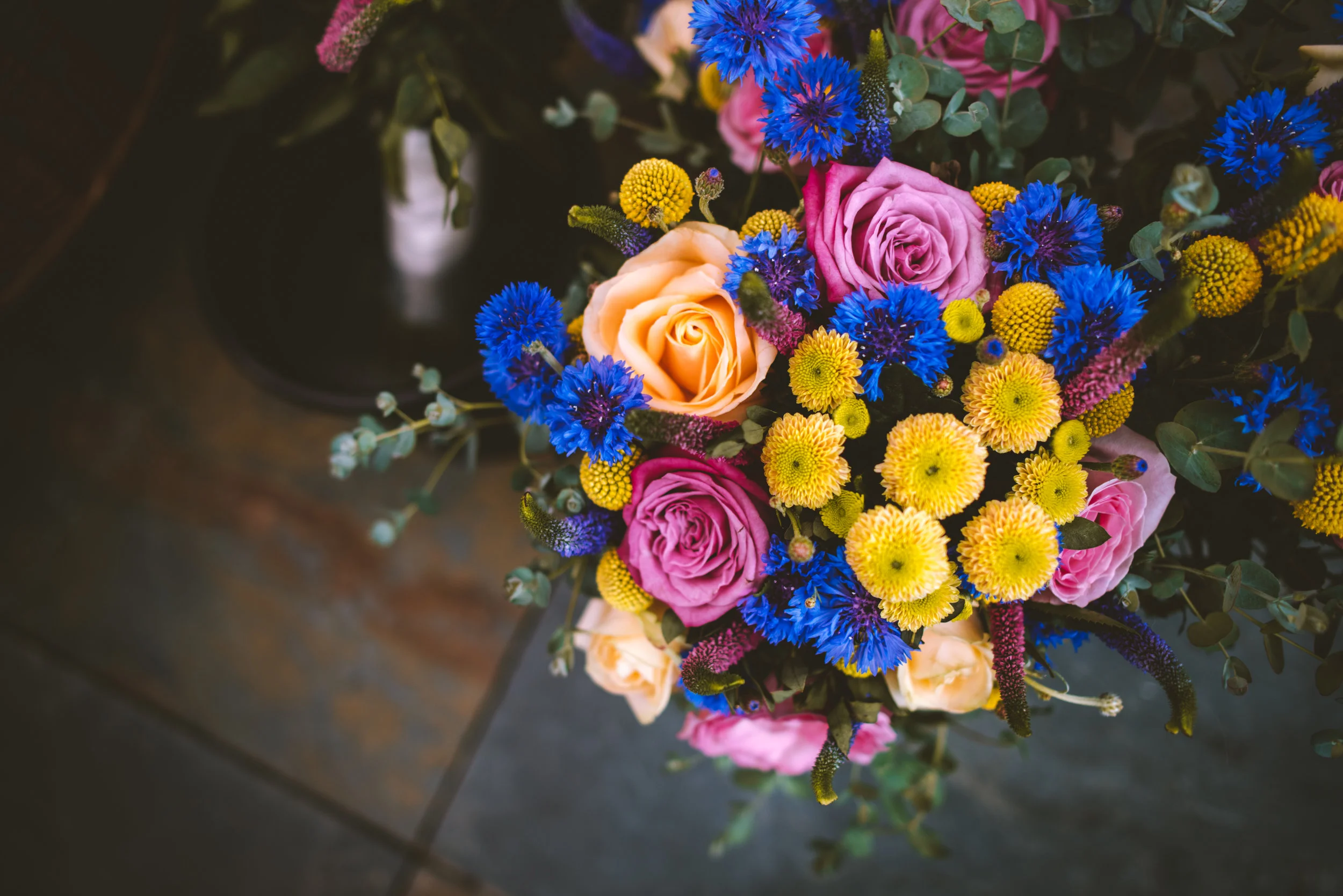 Colorful bouquet of pink and peach roses, yellow billy balls, and bright blue cornflowers with green foliage on a dark surface.
