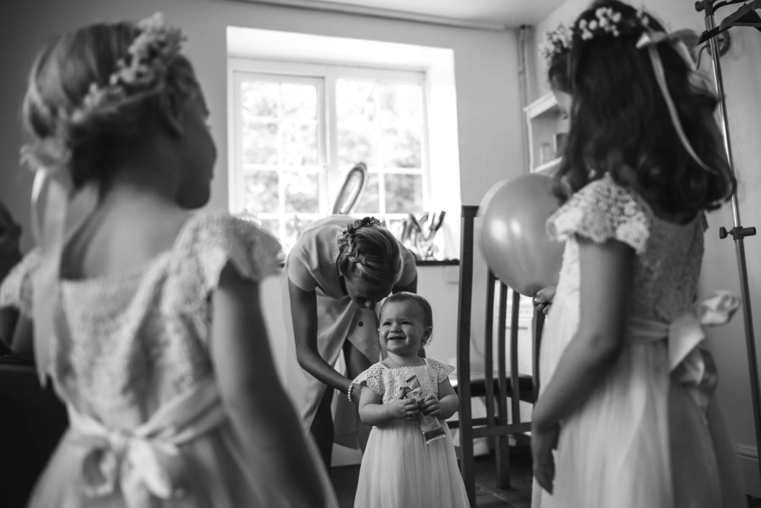 A woman and four young girls in flower girl dresses preparing for a wedding in a room with natural light from a window.