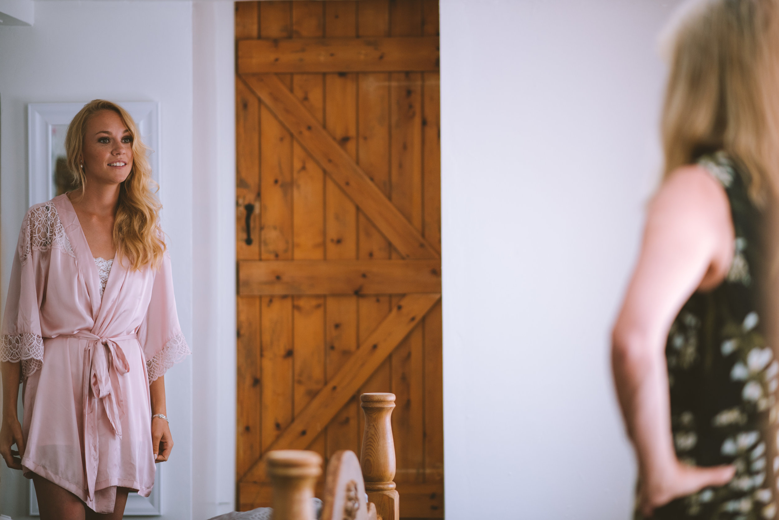 A woman in a pink robe with lace details looking at her reflection in a mirror in a room with wooden accents.