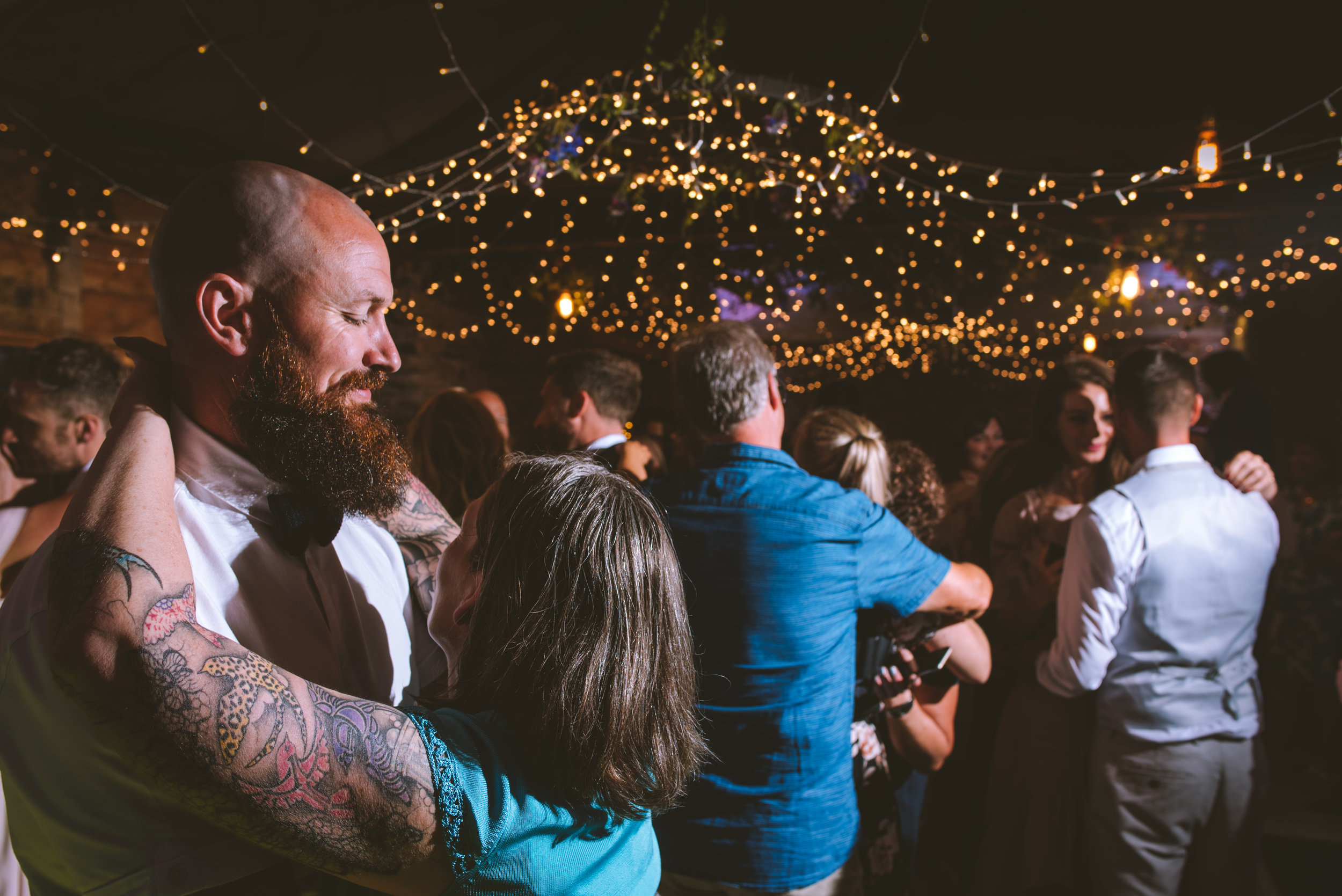 People dancing and socializing at a party or wedding celebration under string lights and a chandelier.